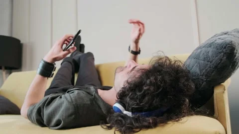 Curly haired guy on the couch listening music with headphones, having fun. Stock Footage 165286857