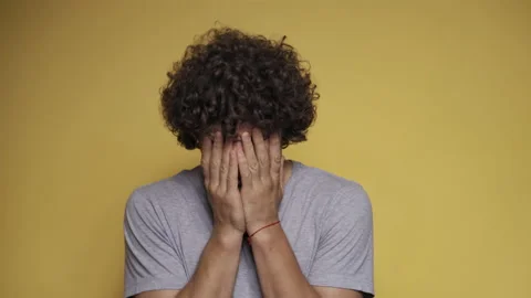 Curly-haired guy sobs with his hands covering his face. Man cries in the studio Stock Footage 140777991