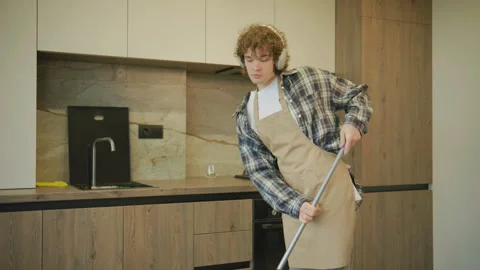 A curly-haired man in a plaid shirt and apron dances lightly while mopping the k Stock Footage 345282353