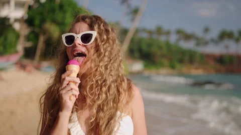 Curly-haired woman eats ice cream on sandy beach. Relishes frosty treat under Stock Footage 270549558