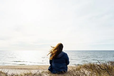 Curly-haired woman in a thin down jacket sitting on the seashore in the evening Stock Photos