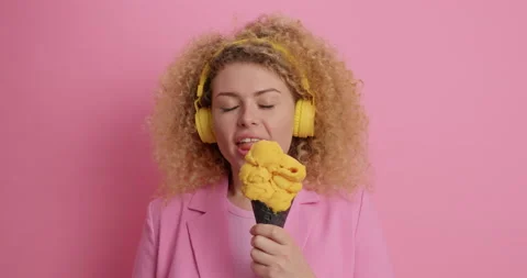 Curly haired young female model licks delicious yellow ice cream wears stereo Stock Footage 154768119