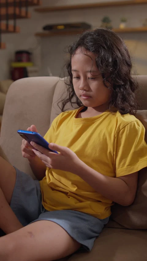 Curly haired young girl using a smartphone while lean back on a living room sofa Stock Footage 339868574
