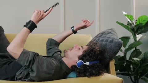 Curly haired young man on the couch listening music with headphones. Stock Footage 154903966