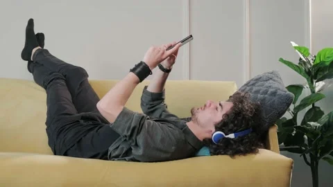 Curly haired young man on the couch listening music with headphones. Stock Footage 155759810