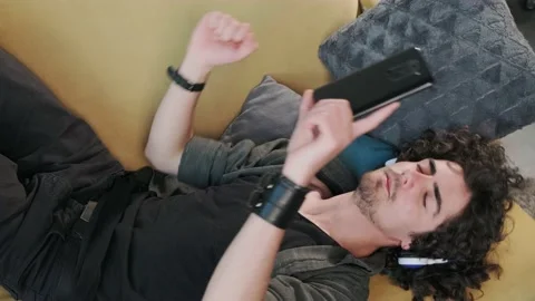 Curly haired young man on the couch listening music with headphones. Stock Footage 156329499