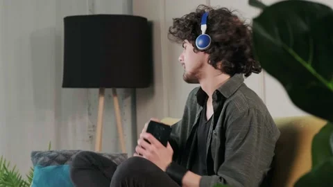 Curly haired young man on the couch listening music with headphones. Stock Footage 158450726