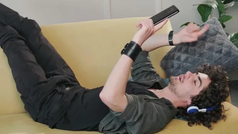 Curly haired young man on the couch listening music with headphones. Stock Footage 165341113