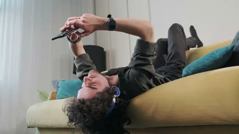 Curly haired young man on the couch listening music with headphones. Stock Footage 168976883