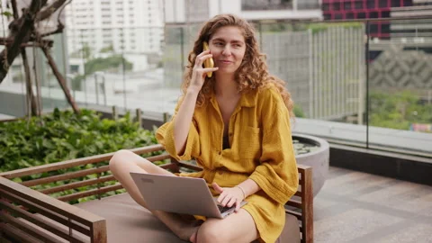 Curly-haired young woman sits on a balcony bench in a big city bakground, Video stock 336695053
