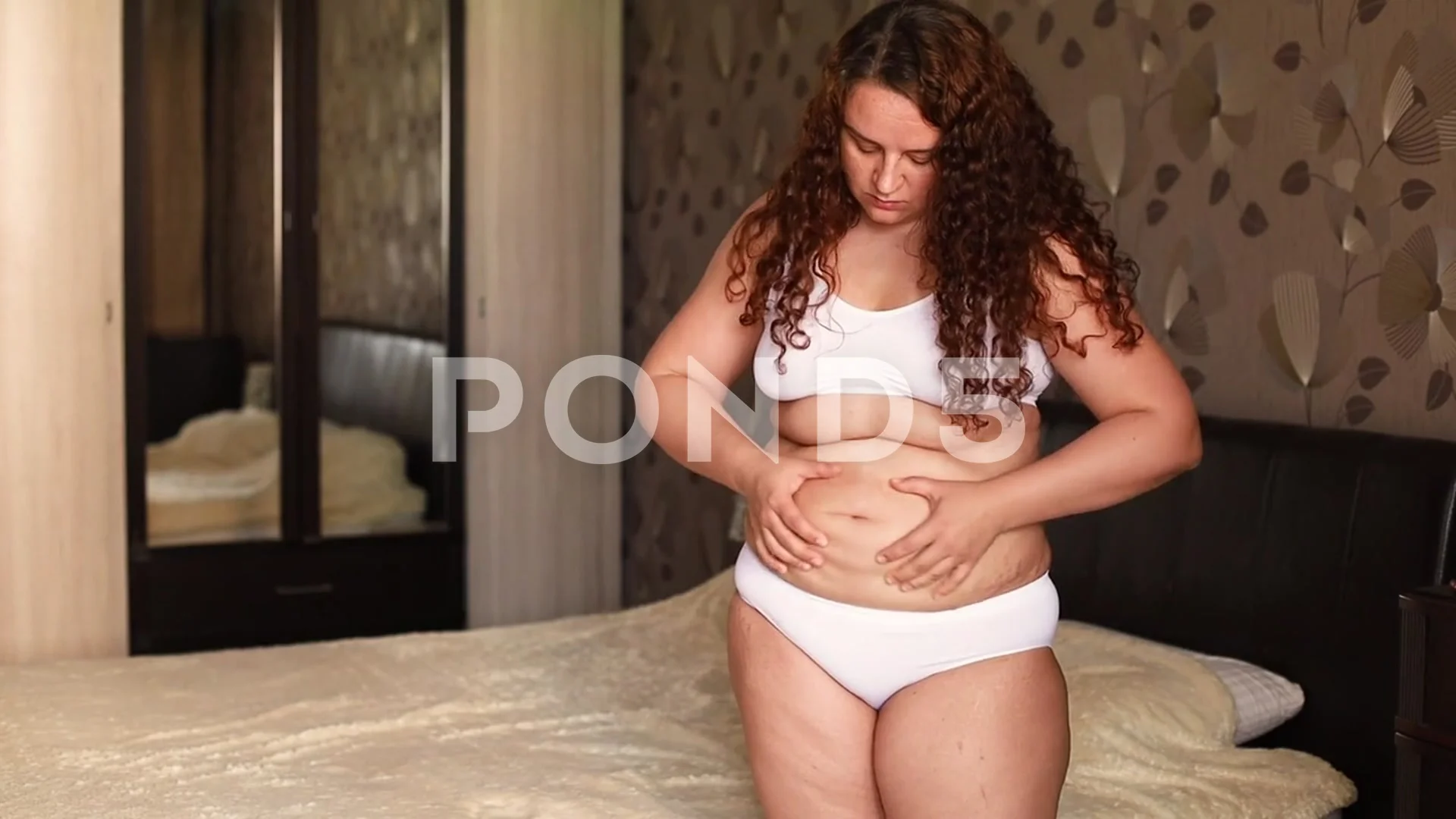 Curly overweight woman in white underwear stand near bed in bedroom, massage