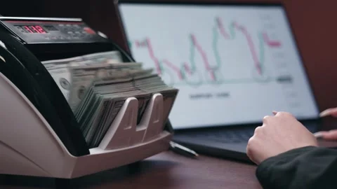Currency counting machine processes banknotes while a laptop displays financial Stock Footage 333979242