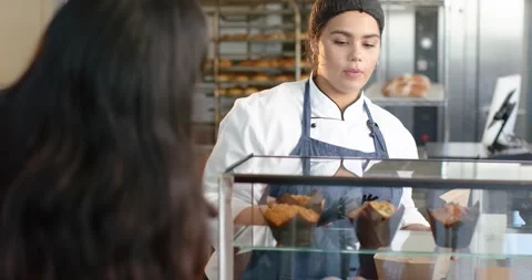 Customer pointing at case, baker in apron grabbing muffin with tongs, packing Stock Footage 337933320