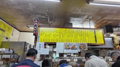 Customers view a yellow menu at a busy Japanese food stall Stock Footage 346011785