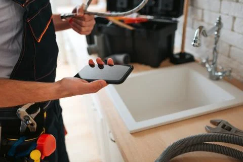 Cut view of man standing in kitchen at sink. He hold phone and wrench. Hose on Фото