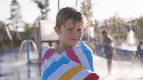 Cute 6 year old boy wrapped in towel smiles in front of splash pad, sunset Stock Footage 233940516
