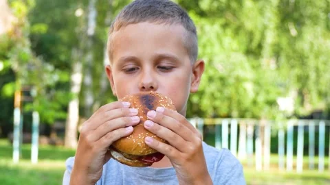 Cute 9-year-old boy enjoys eating a hamburger in a city park in the summer Stock Footage 247041648