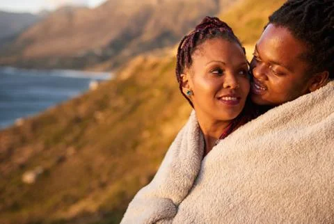 Cute affectionate interracial couple keeping warm with a blanket outdoors Fotos Stock