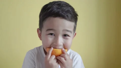 Cute asian boy eats a piece of orange on colour background Stock Footage 195038236