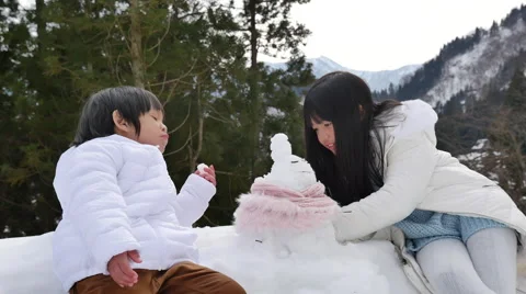 Cute asian sibling in winter clothes making a little snowman with old village Stock Footage
