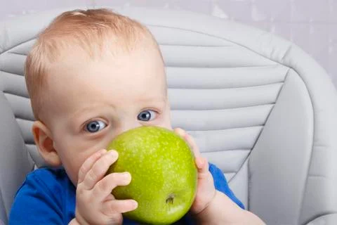 Cute baby 1,4 years old sitting on high children chair and eating green apple Stock Photos