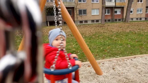 A cute baby boy in a red jacket and a blue hat on a chain swing at a park. 4K Stock Footage 143909443