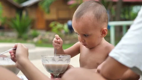 Cute baby eats breakfast from cup himself with spoon smeared with food. Feeding Stock Footage 228706043