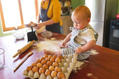 Cute baby playing with egg carton on wooden table while mother preparing food in Stock Photos