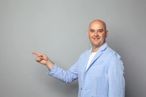 Cute bald man in a jacket and t-shirt shows against a gray wall Foto stock