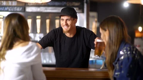 Cute barman  serving beer to a couple of pretty girls waiting at the bar tab Stock Footage