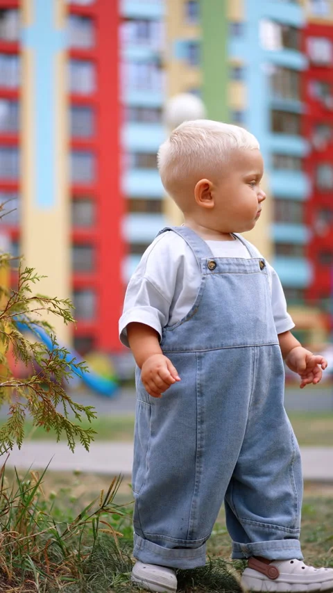 Cute blond baby boy in jeans romper stands at the bush