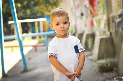 A cute blond boy in a white T-shirt stands modestly and looks into the lens with Stock Photos