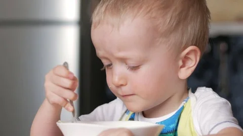 A cute blonde boy eats a porridge with a spoon from a plate of the house. Close Video stock 75043790