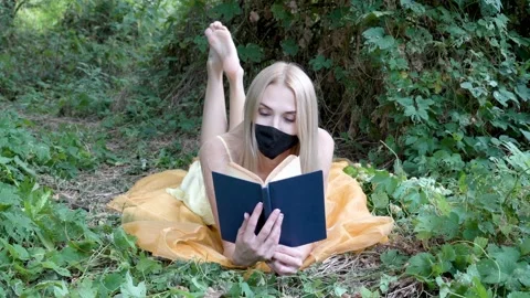 Cute blonde in a protective mask is lying and reading a book on the grass in Video stock 141863933