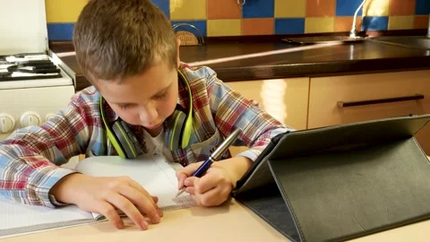 A cute boy of 6-7-8 years old looks at the tablet, answers and writes something Stock Footage 230993580