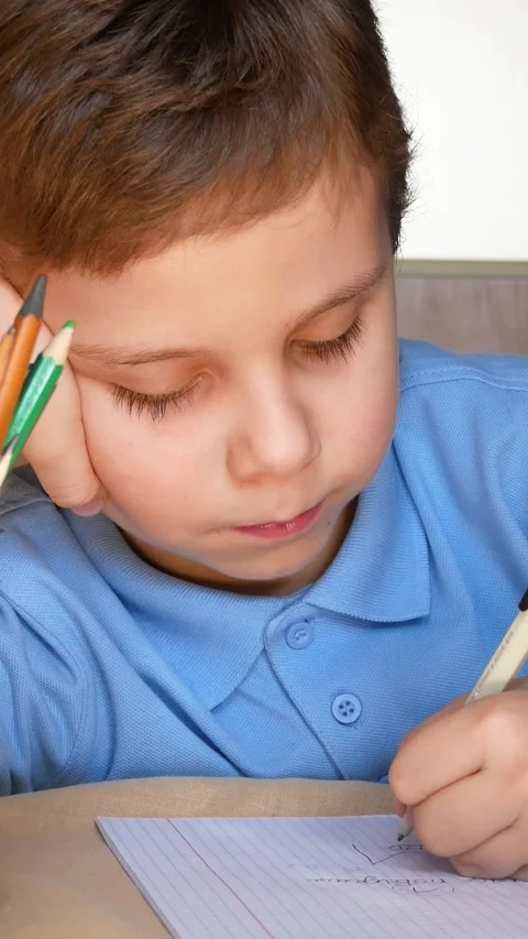 A cute boy of 6-7 years old writes with a pen in a notebook, resting his head. Video stock 317849355