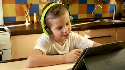 A cute boy of 7-8 years old in headphones sits in front of a tablet and talks to Stock Footage 230661539