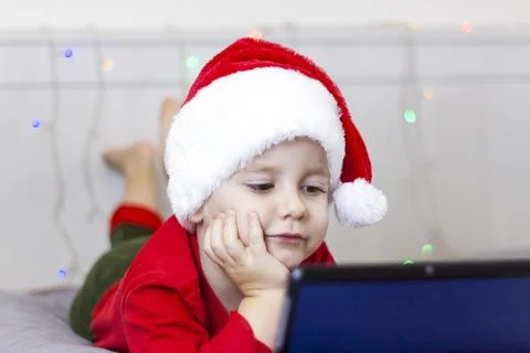 A cute boy is in bed looking at a tablet. Funny smiling child. Wearing a Sant Stock Photos