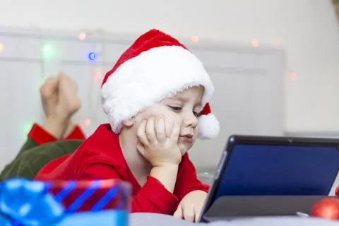 A cute boy is in bed looking at a tablet. Funny smiling child. Wearing a Sant Stock Photos