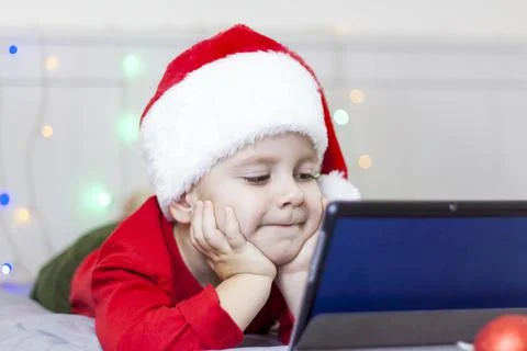 A cute boy is in bed looking at a tablet. Funny smiling child. Wearing a Sant Stock Photos