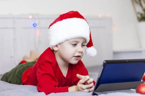 A cute boy is in bed looking at a tablet. Funny smiling child. Wearing a Sant Stock Photos