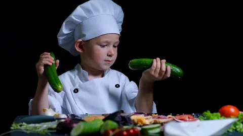 Cute boy bites a cucumber while cooking. He is wearing chefs suit ans cap. Black Stock Footage 92562685