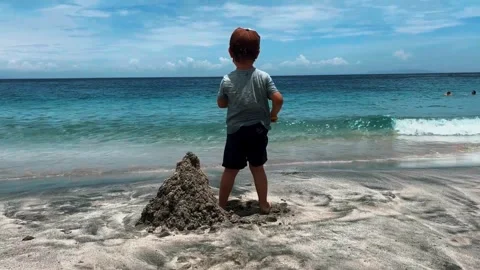 A cute boy is building a sand castle on the shore of the blue sea on a hot sunny Vídeos de archivo 264992021