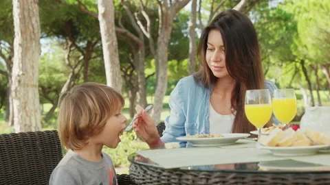 Cute boy enjoys his breakfast and fruity drink, while sitting by his mom Stock Footage 163243701