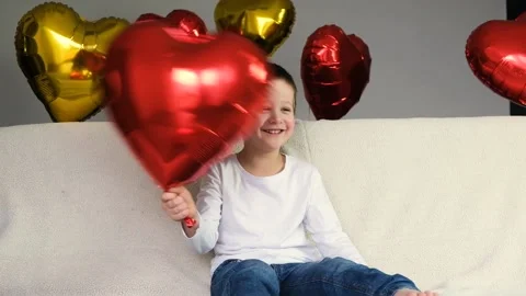 Cute boy is indulging with a red heart-shaped balloon on a light sofa Stock Footage 146164542