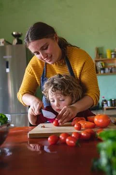 Cute boy learning to cut fresh tomatoes with knife from mother at kitchen Stock Photos