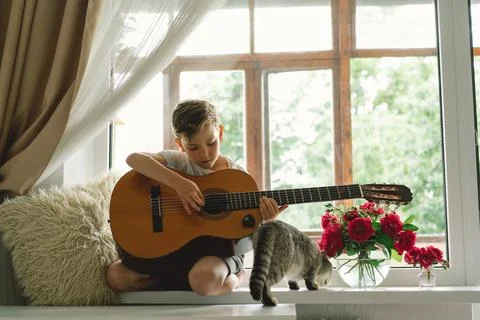 Cute boy learns to play the classical guitar on the windowsill near the window. Stock Photos