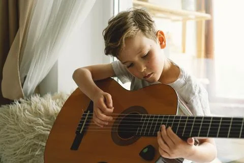 Cute boy learns to play the classical guitar on the windowsill near the window. Stock Photos
