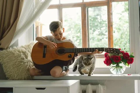 Cute boy learns to play the classical guitar on the windowsill near the window. Stock Photos
