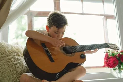 Cute boy learns to play the classical guitar on the windowsill near the window. Stock Photos